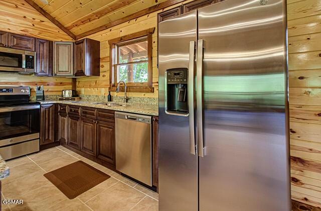 kitchen with light stone countertops, sink, stainless steel appliances, wood walls, and light tile patterned floors