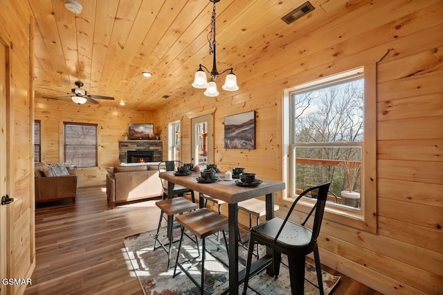 dining space with a stone fireplace, wood-type flooring, wooden walls, wood ceiling, and ceiling fan with notable chandelier