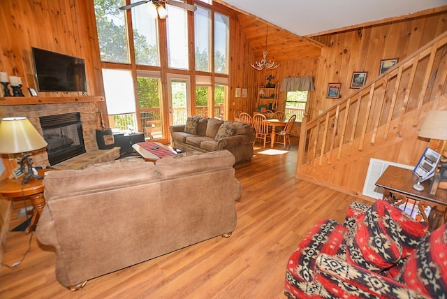 living room with ceiling fan with notable chandelier, hardwood / wood-style flooring, a high ceiling, a stone fireplace, and wood walls