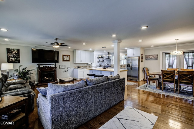 living area featuring dark wood-style floors, decorative columns, a warm lit fireplace, ceiling fan, and ornamental molding