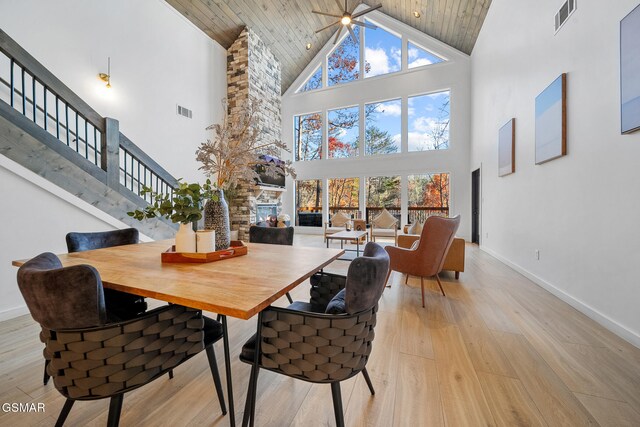 dining space featuring a stone fireplace, ceiling fan, a towering ceiling, light wood-type flooring, and wood ceiling