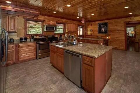 kitchen with appliances with stainless steel finishes, wood ceiling, an island with sink, sink, and wood walls