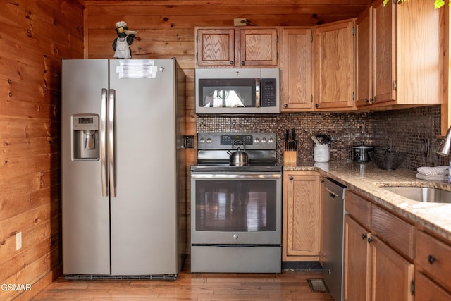 kitchen with appliances with stainless steel finishes, wooden walls, sink, light stone counters, and light hardwood / wood-style flooring