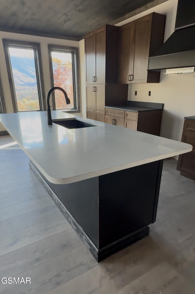 kitchen with wall chimney exhaust hood, dark brown cabinetry, a kitchen island with sink, sink, and light hardwood / wood-style flooring