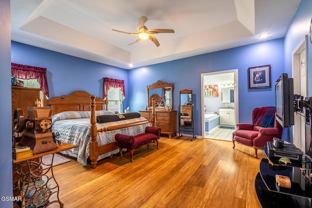 bedroom with a tray ceiling, light wood-type flooring, ensuite bath, and a ceiling fan
