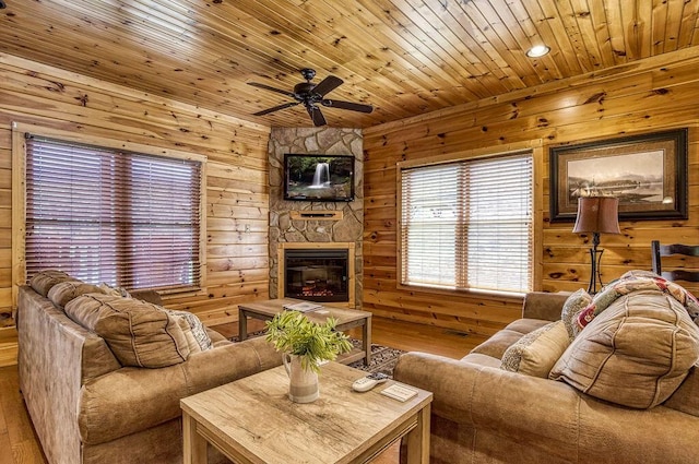 living room featuring ceiling fan, wood walls, a stone fireplace, and wooden ceiling