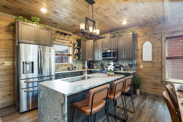 kitchen with light stone countertops, wooden ceiling, an island with sink, decorative light fixtures, and appliances with stainless steel finishes