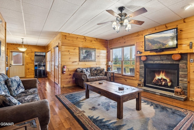 living room featuring a stone fireplace, wood finished floors, a ceiling fan, and wooden walls