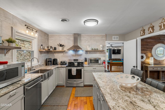 kitchen featuring sink, appliances with stainless steel finishes, light stone countertops, wall chimney exhaust hood, and light wood-type flooring