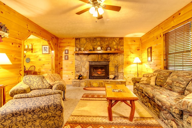 carpeted living room featuring ceiling fan, a stone fireplace, a textured ceiling, and wooden walls