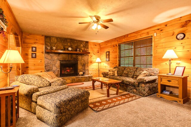 living room with a textured ceiling, light colored carpet, ceiling fan, wooden walls, and a stone fireplace