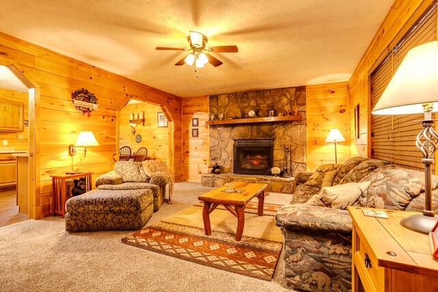 living room featuring carpet, wood walls, a stone fireplace, ceiling fan, and a textured ceiling