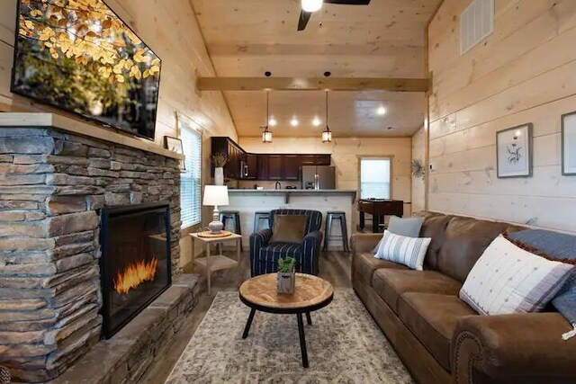 living room with vaulted ceiling with beams, a stone fireplace, ceiling fan, and wooden walls