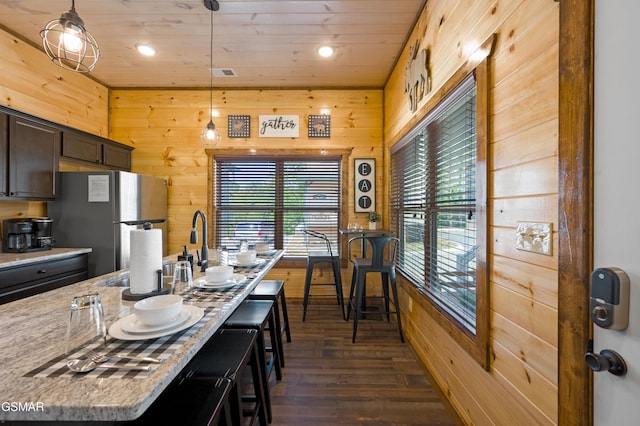 kitchen with decorative light fixtures, wooden walls, light stone countertops, and stainless steel refrigerator