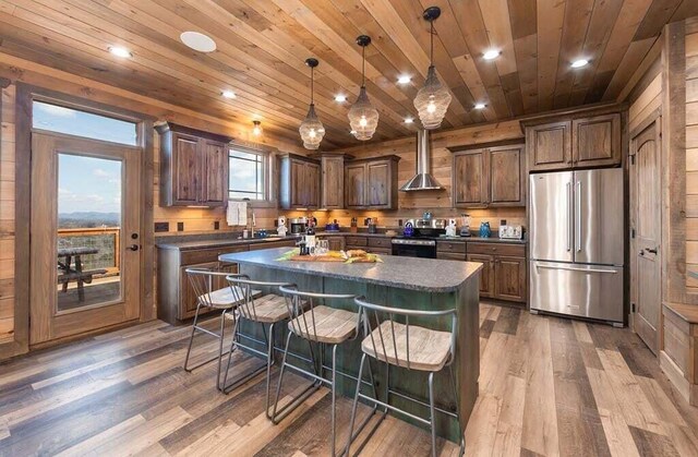 kitchen featuring wall chimney exhaust hood, stainless steel appliances, decorative light fixtures, hardwood / wood-style floors, and a center island