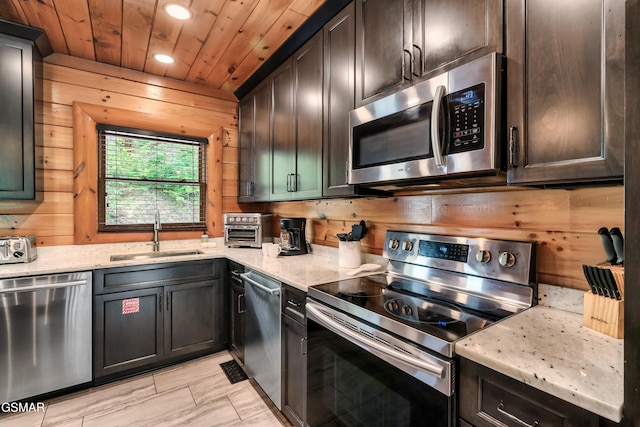 kitchen with light stone countertops, appliances with stainless steel finishes, dark brown cabinets, wooden walls, and sink