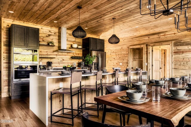 kitchen featuring ventilation hood, an island with sink, wooden walls, wood ceiling, and appliances with stainless steel finishes