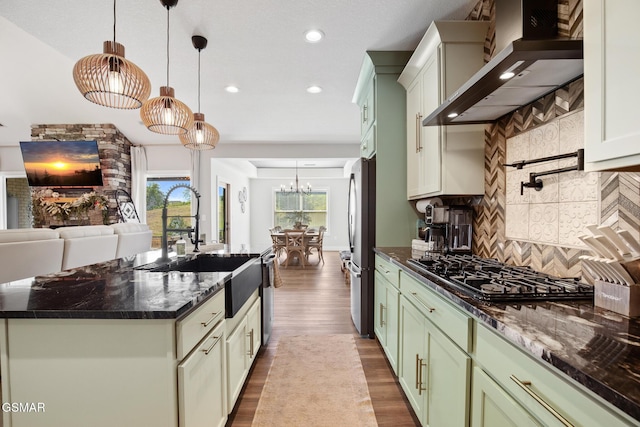 kitchen featuring stainless steel appliances, wall chimney range hood, an island with sink, dark stone countertops, and pendant lighting