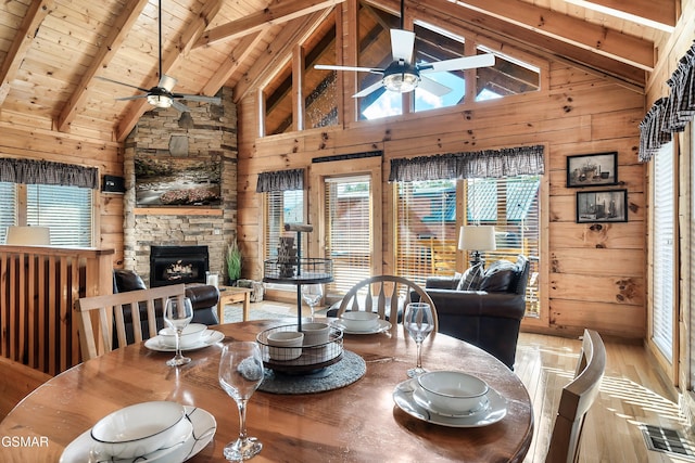 dining area featuring a stone fireplace, wood walls, ceiling fan, wooden ceiling, and beam ceiling