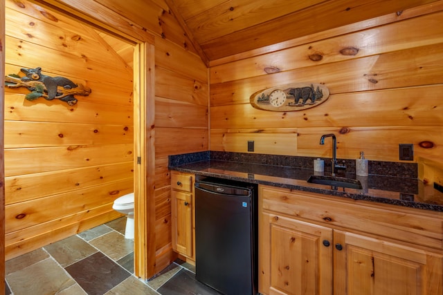 kitchen with stone tile floors, wooden walls, a sink, vaulted ceiling, and dark stone counters