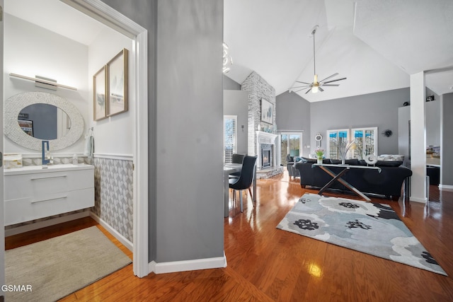 interior space with a wainscoted wall, ceiling fan, wood finished floors, a stone fireplace, and high vaulted ceiling