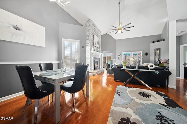 dining room with a large fireplace, ceiling fan, wood finished floors, high vaulted ceiling, and baseboards