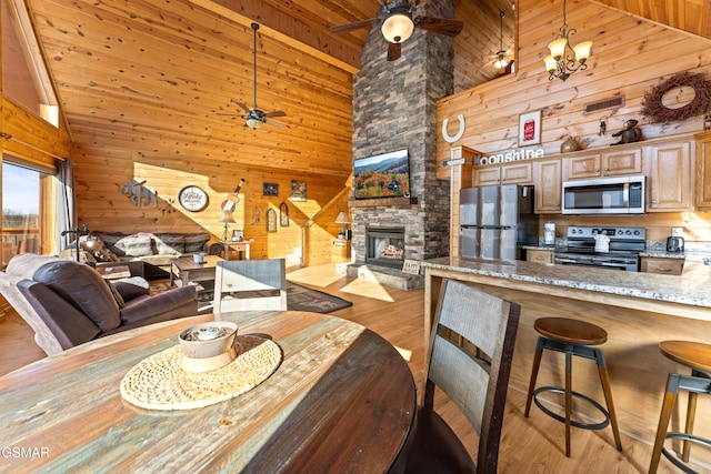 dining room with wood walls, high vaulted ceiling, light wood-type flooring, a fireplace, and ceiling fan with notable chandelier