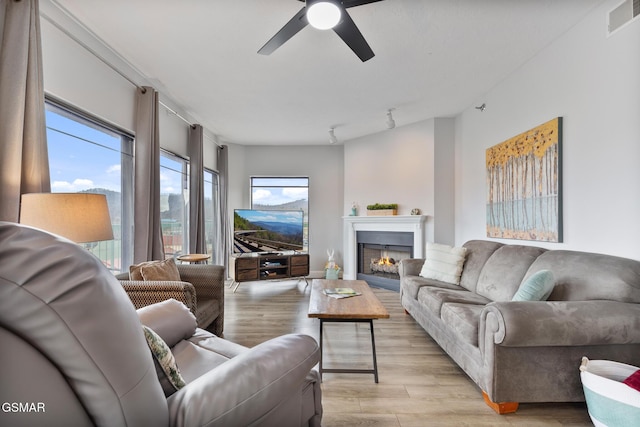 living room featuring a warm lit fireplace, light wood-style flooring, visible vents, and a ceiling fan