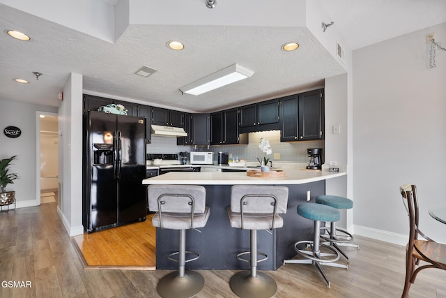 kitchen featuring white microwave, under cabinet range hood, a peninsula, black fridge with ice dispenser, and range