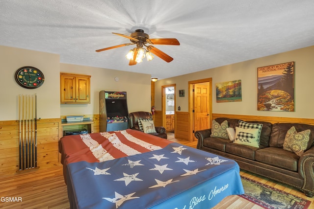 bedroom with a textured ceiling, wood walls, light wood-style flooring, a wainscoted wall, and a ceiling fan