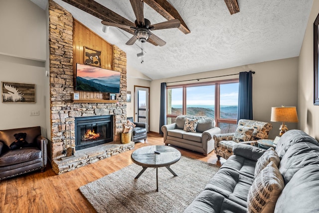 living room with ceiling fan, vaulted ceiling with beams, a stone fireplace, light hardwood / wood-style floors, and a textured ceiling