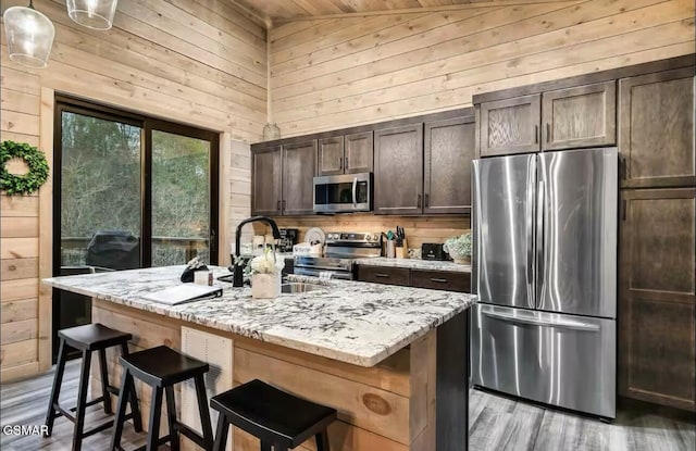 kitchen with wood walls, a kitchen island with sink, appliances with stainless steel finishes, dark brown cabinetry, and a sink
