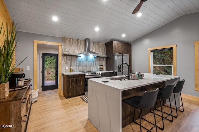 kitchen featuring wall chimney exhaust hood, lofted ceiling, appliances with stainless steel finishes, a sink, and a kitchen breakfast bar