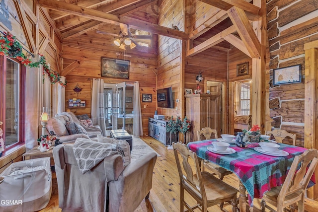 dining space featuring ceiling fan, light hardwood / wood-style flooring, beamed ceiling, high vaulted ceiling, and wooden walls