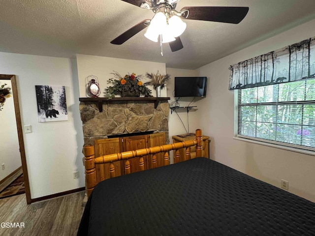 bedroom featuring a stone fireplace, a textured ceiling, ceiling fan, and wood finished floors