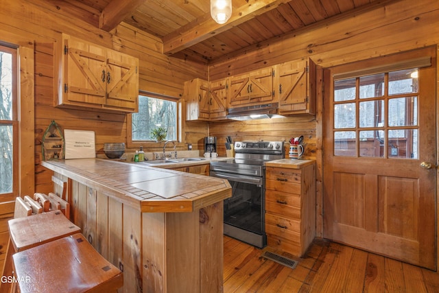 kitchen featuring tile countertops, a peninsula, stainless steel range with electric cooktop, under cabinet range hood, and a sink