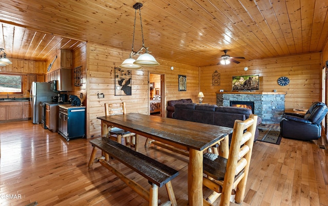 dining room with light wood-style flooring, a ceiling fan, wood ceiling, wood walls, and a stone fireplace