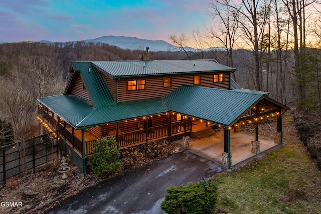 back of property at dusk with a forest view, covered porch, metal roof, a mountain view, and log veneer siding