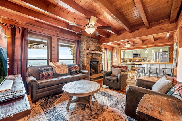 living room with wooden ceiling, a stone fireplace, a water view, ceiling fan, and beam ceiling