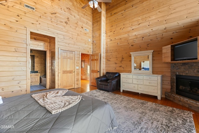 bedroom with a stone fireplace, ensuite bath, wooden walls, light hardwood / wood-style floors, and a high ceiling