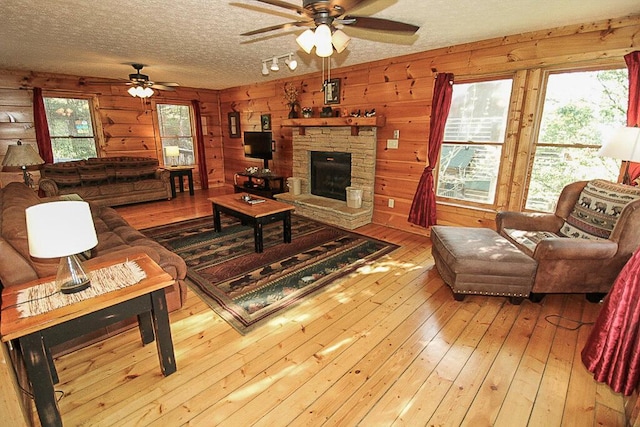 living room featuring light wood-type flooring, a textured ceiling, ceiling fan, a stone fireplace, and wood walls