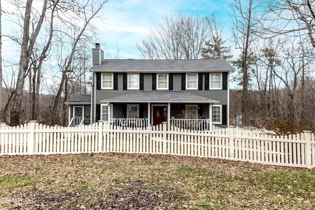 colonial-style house featuring covered porch, roof with shingles, a chimney, and a fenced front yard