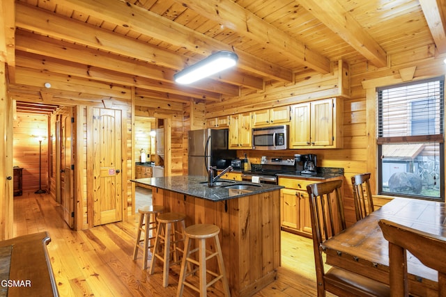 kitchen with stainless steel appliances, a kitchen island with sink, wooden walls, sink, and beamed ceiling