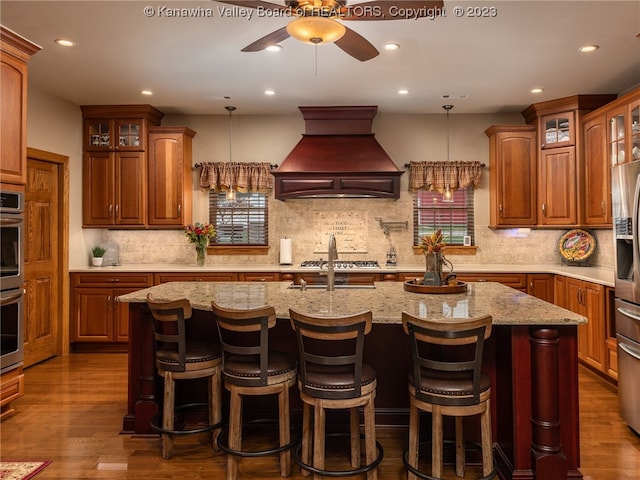 kitchen featuring dark wood-type flooring, custom range hood, an island with sink, backsplash, and ceiling fan