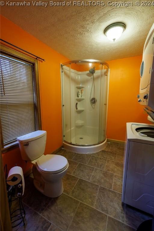 bathroom with stacked washer / drying machine, a shower with door, toilet, and a textured ceiling