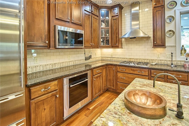 kitchen featuring appliances with stainless steel finishes, sink, light wood-type flooring, wall chimney exhaust hood, and light stone counters