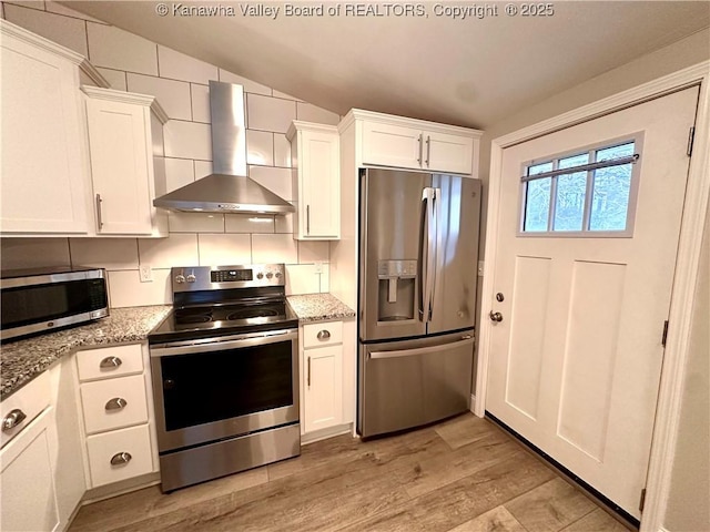 kitchen featuring wall chimney range hood, decorative backsplash, light stone countertops, appliances with stainless steel finishes, and white cabinetry