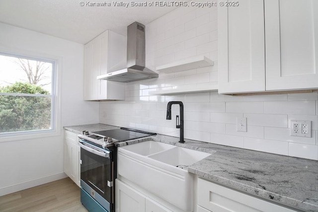 kitchen featuring light wood-type flooring, light stone counters, stainless steel range with electric stovetop, wall chimney exhaust hood, and white cabinets