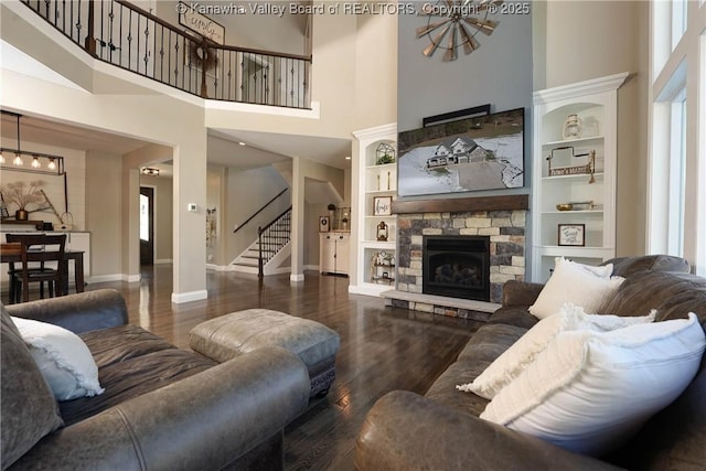 living room featuring built in shelves, a stone fireplace, dark wood-type flooring, and a towering ceiling