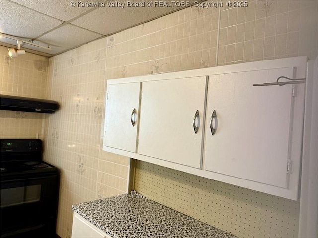 kitchen featuring black electric range oven, a paneled ceiling, tile walls, light stone counters, and exhaust hood